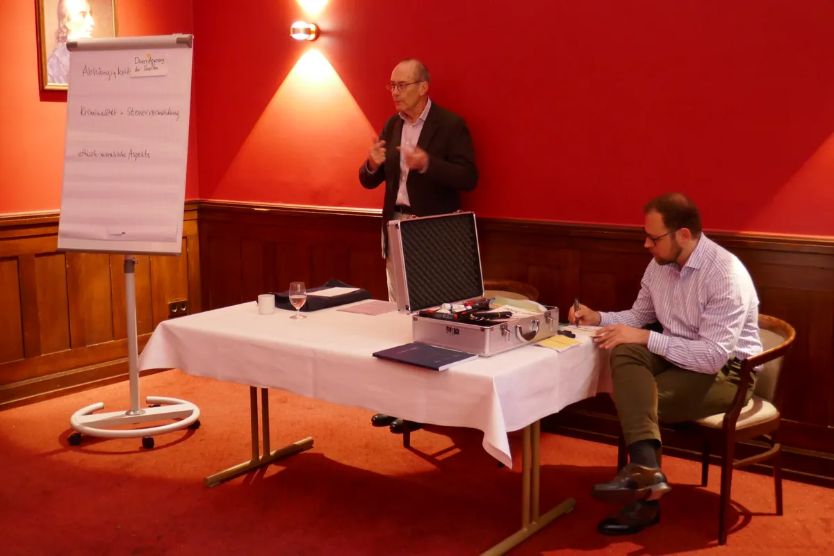 Hermann-Ehlers-Symposion 2022 in Hamburg: Ein Referent erläutert am Flipchart Stichworte wie Abhängigkeit und Steuervermeidung, ein Teilnehmer macht sich Notizen